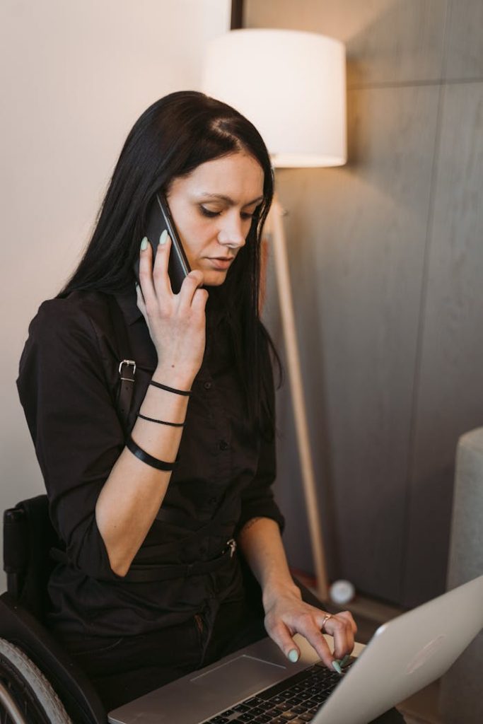 A woman in a wheelchair multitasks with a smartphone and laptop indoors. Ideal for remote work themes.