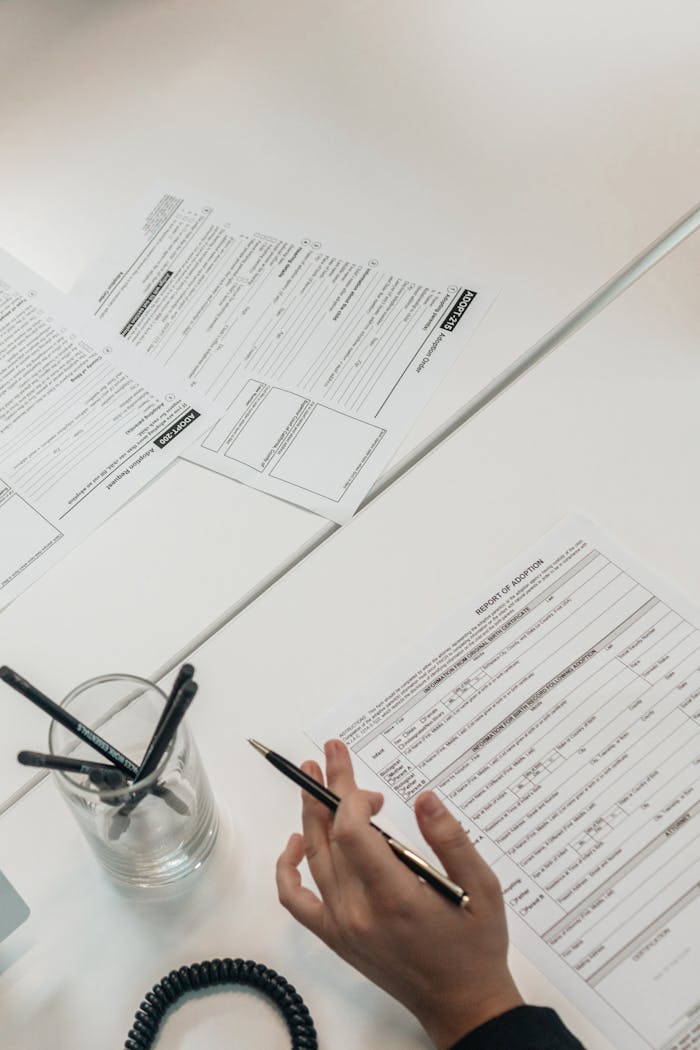 A person holds a pen while examining business documents on a white desk, top view.